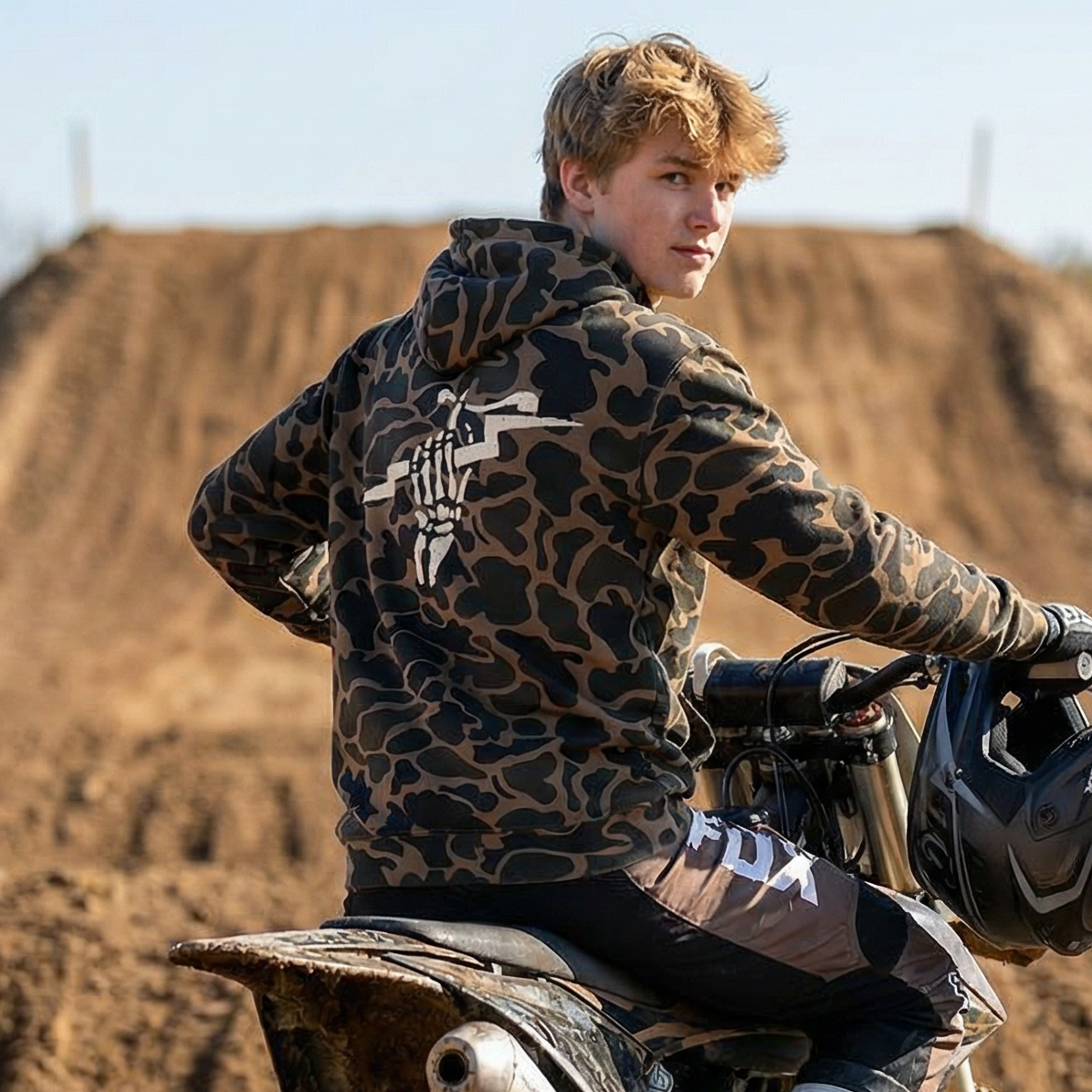 Teen male sitting on his dirt bike in the middle of a motocross track wearing the basic moto 'fist of lightning' vintage duck camo hoodie.
